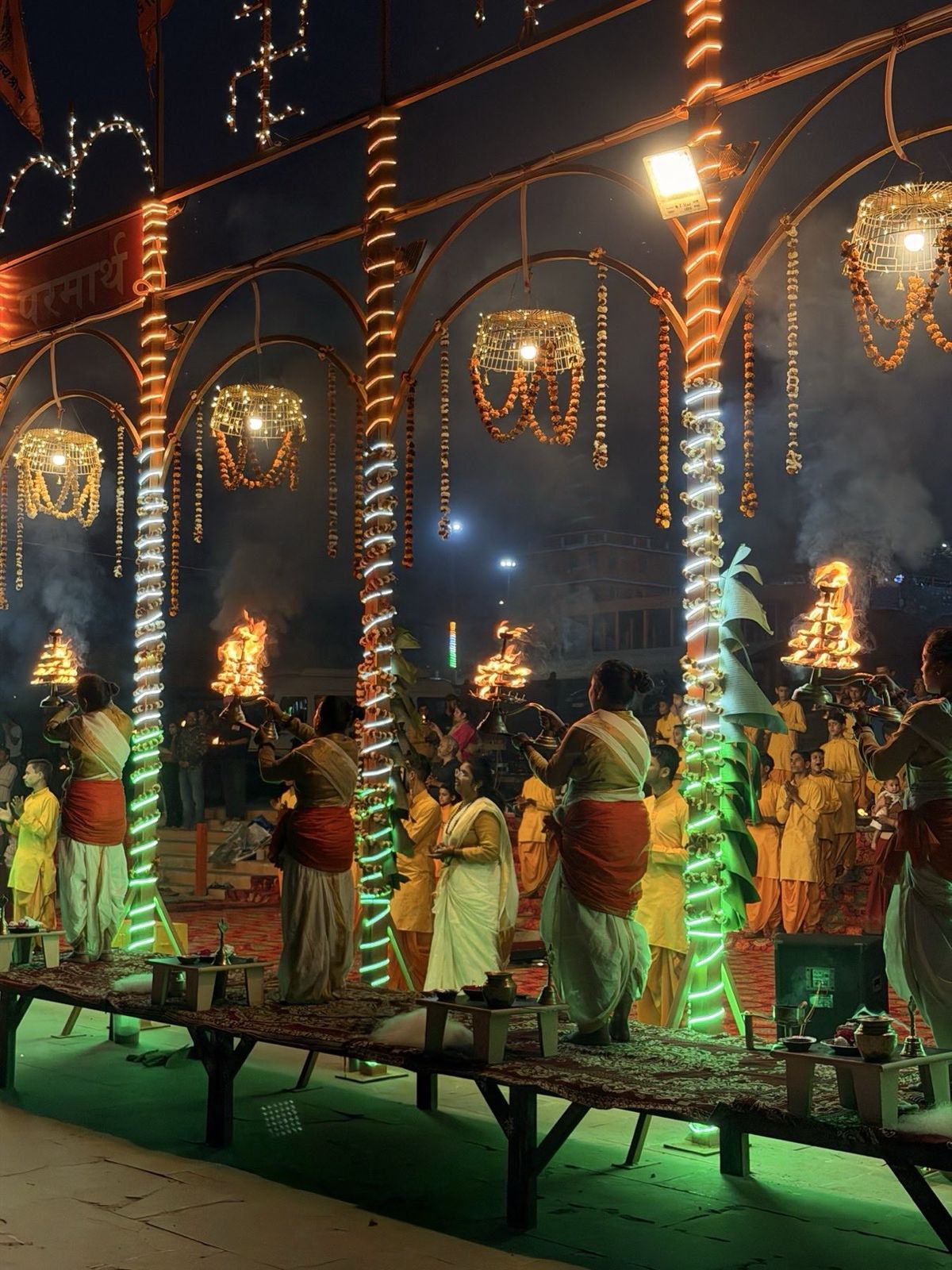 Priests lifting flaming aarti lamps at an evening ceremony by the river, hung with marigolds and lights